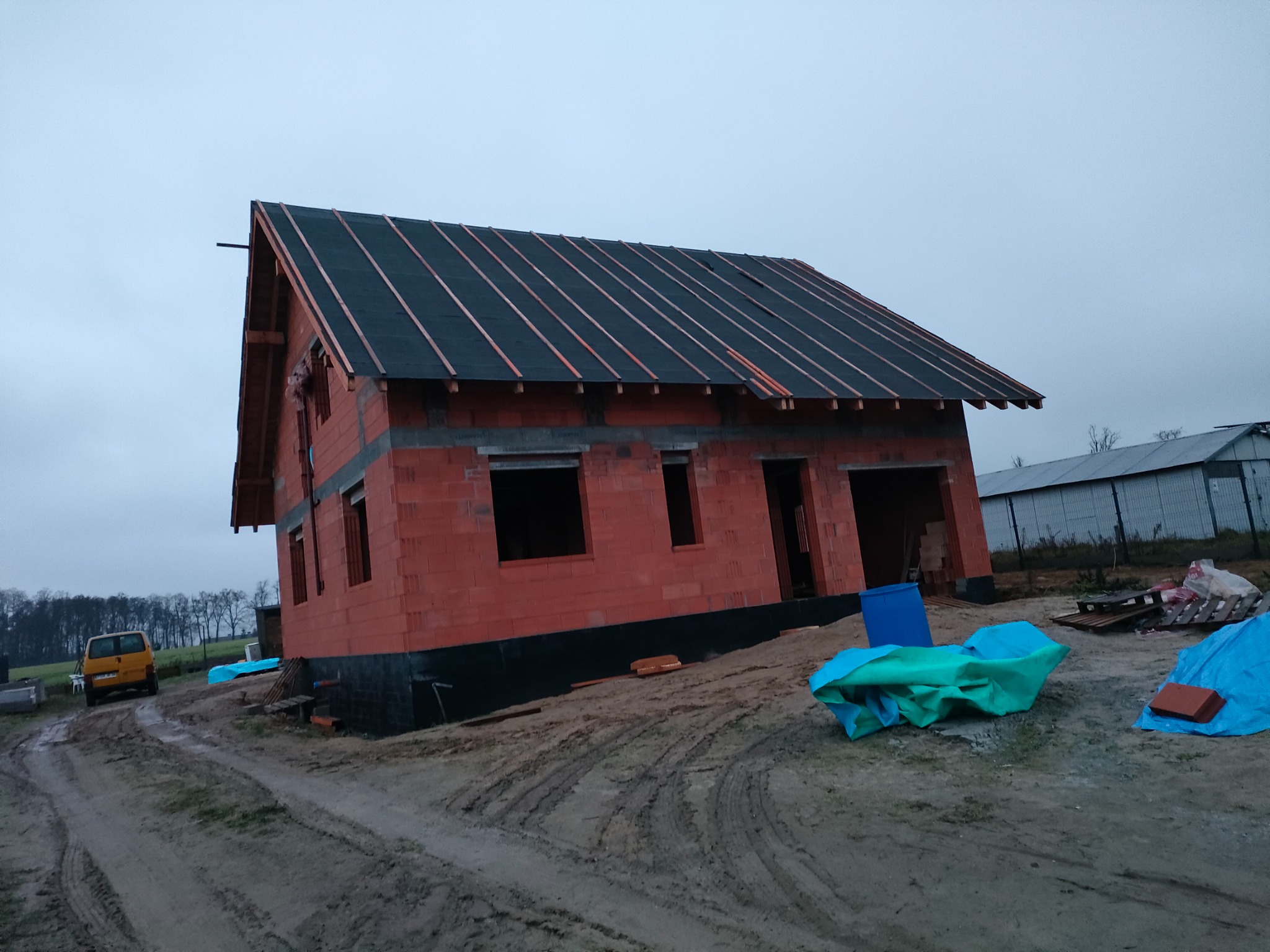 Surowy stan domu murowanego z czerwonej cegły z częściowo wykończonym czarnym dachem z łatami, otoczonego błotnistym terenem i elementami budowlanymi.