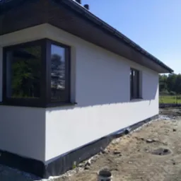 Boczny widok otynkowanego, parterowego domu z brązowymi oknami i dachem, z widoczną hydroizolacją przy gruncie. Jasne, słoneczne ujęcie.