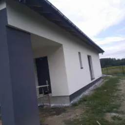 Nowoczesny dom jednorodzinny po remoncie elewacji. Jasna fasada z ciemnym akcentem na kolumnie wejściowej. Widoczne rusztowanie.