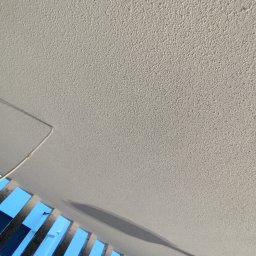 Kamil Wojtanek - Teksturowana, jasna ściana z widoczną listwą sufitową i fragmentem balustrady z metalowymi szczeblami na tle niebieskiego nieba.