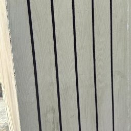 Kamil Wojtanek - Jasnoszara elewacja z pionowych desek z ciemnymi fugami, imitująca drewno. Widoczny fragment narożnika budynku w słoneczny dzień.