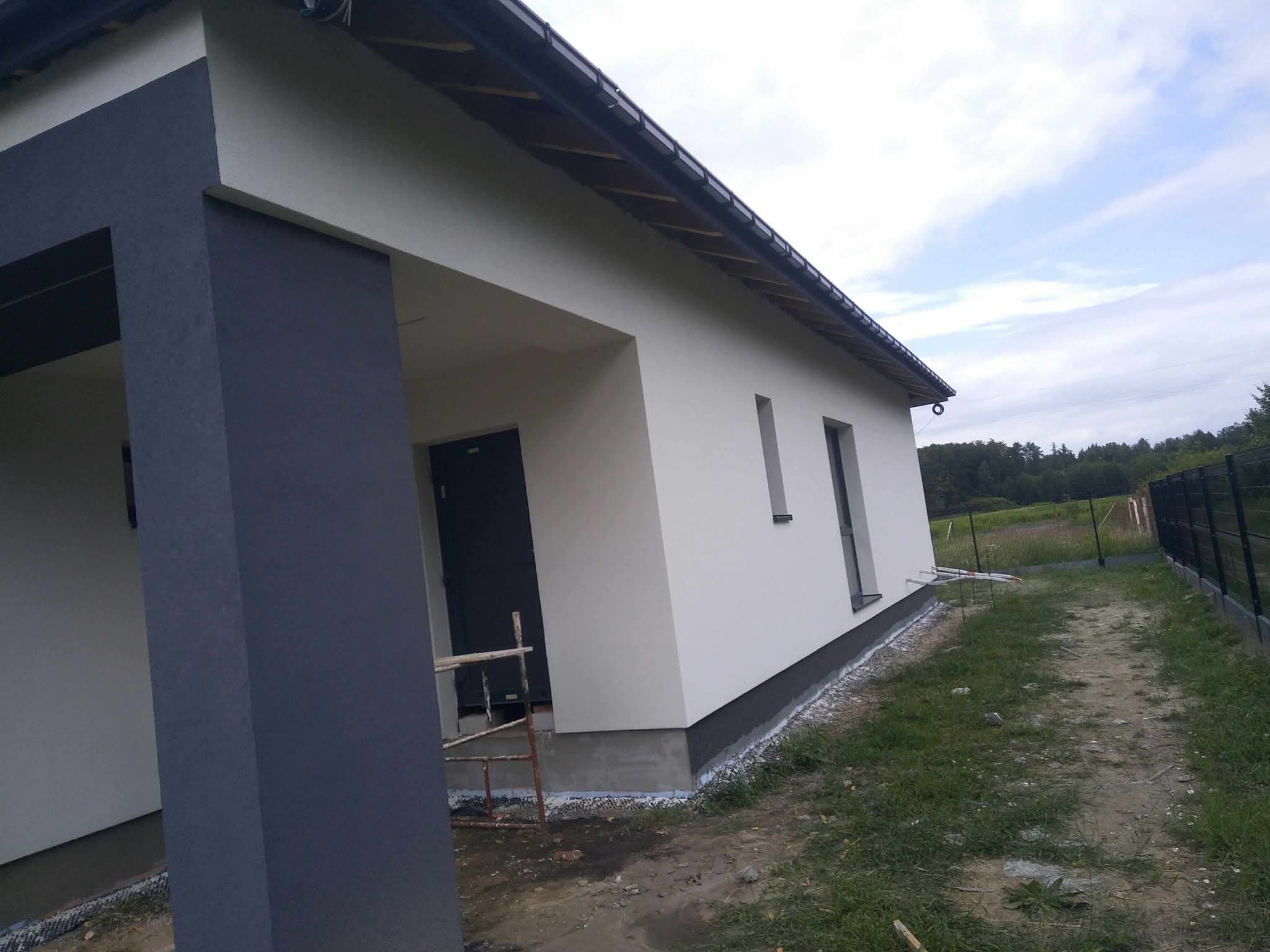 Nowoczesny dom jednorodzinny po remoncie elewacji. Jasna fasada z ciemnym akcentem na kolumnie wejściowej. Widoczne rusztowanie.