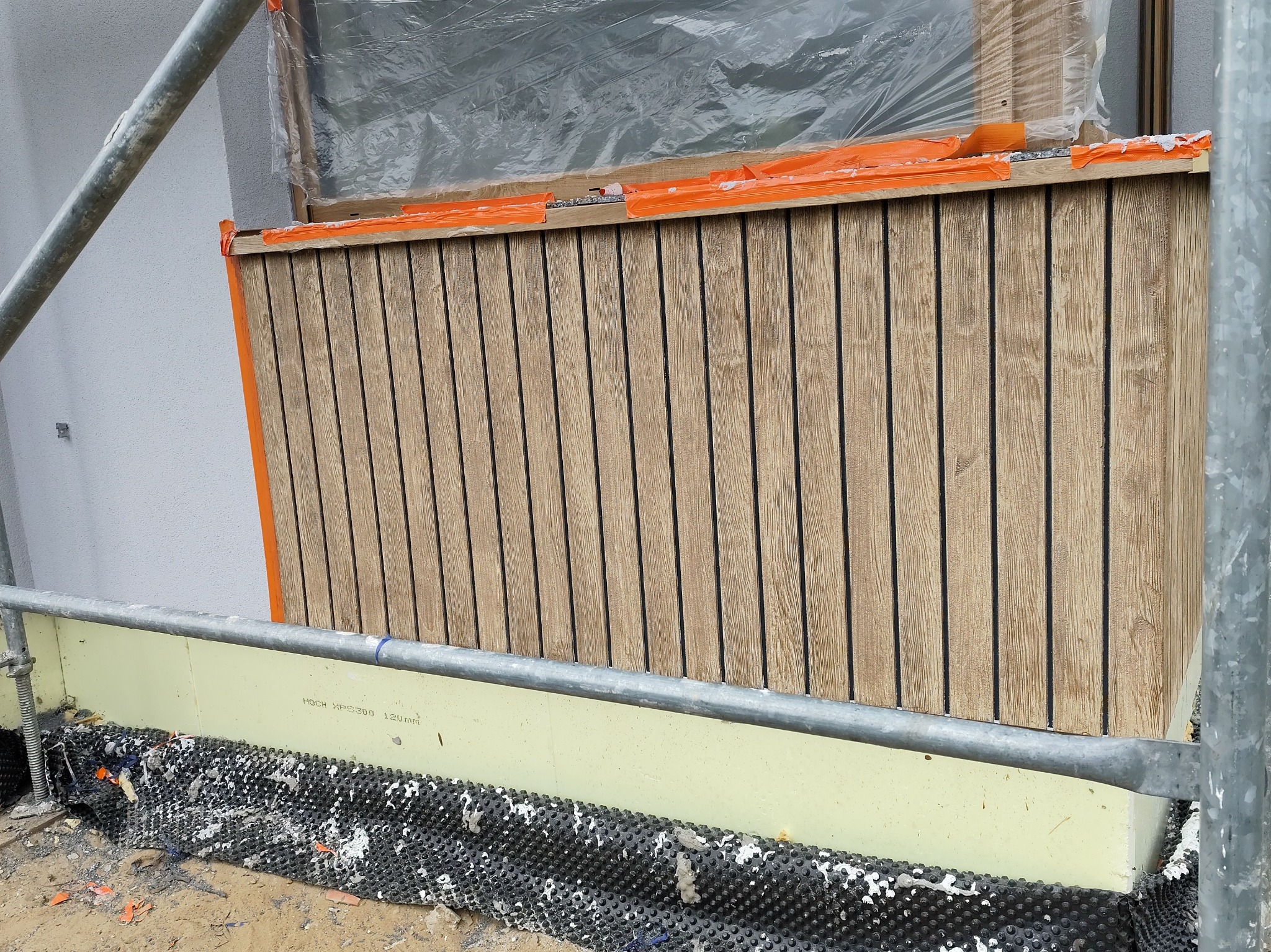 Montaż drewnianej elewacji na ocieplonym budynku. Widoczne rusztowanie i zabezpieczenia okna folią. Deski ułożone pionowo, z widoczną strukturą drewna.