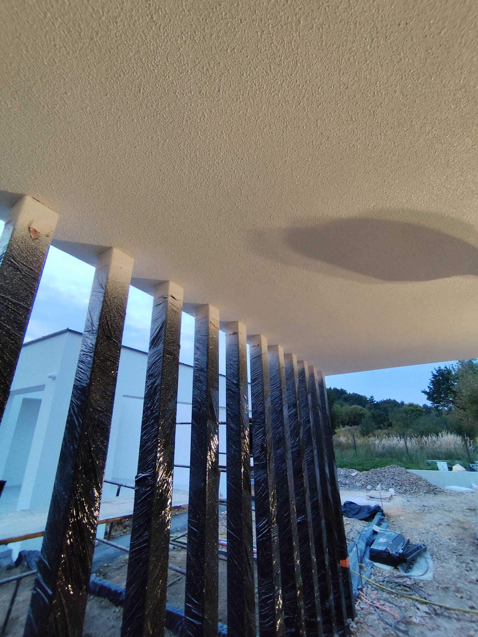 Nowoczesna elewacja domu w trakcie budowy z widocznymi czarnymi, pionowymi elementami dekoracyjnymi i surowym wykończeniem sufitu. Widok z dołu, plac budowy w tle.