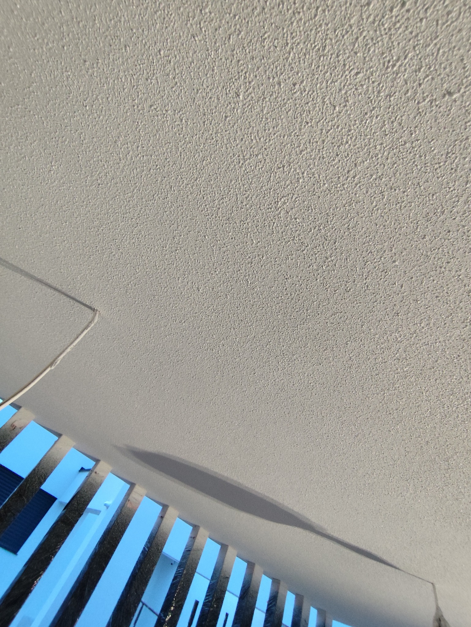 Teksturowana, jasna ściana z widoczną listwą sufitową i fragmentem balustrady z metalowymi szczeblami na tle niebieskiego nieba.