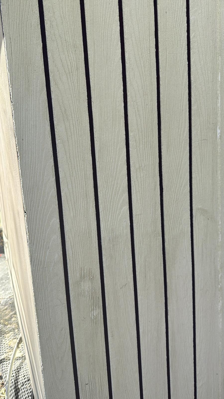 Jasnoszara elewacja z pionowych desek z ciemnymi fugami, imitująca drewno. Widoczny fragment narożnika budynku w słoneczny dzień.