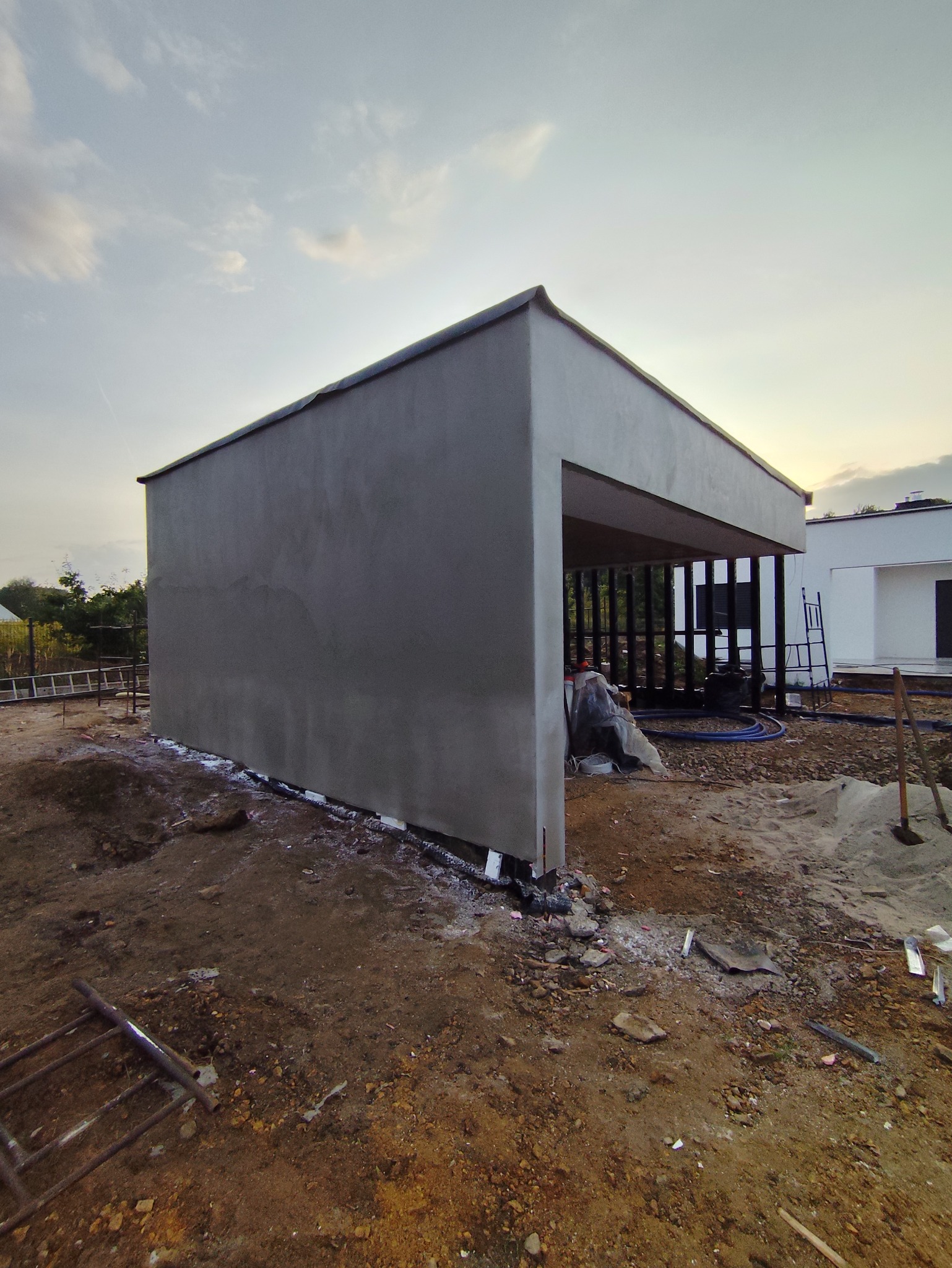 Surowy, szary budynek w trakcie prac wykończeniowych z widocznymi rusztowaniami i narzędziami. Tło stanowi niebo o zmierzchu i sąsiedni budynek.