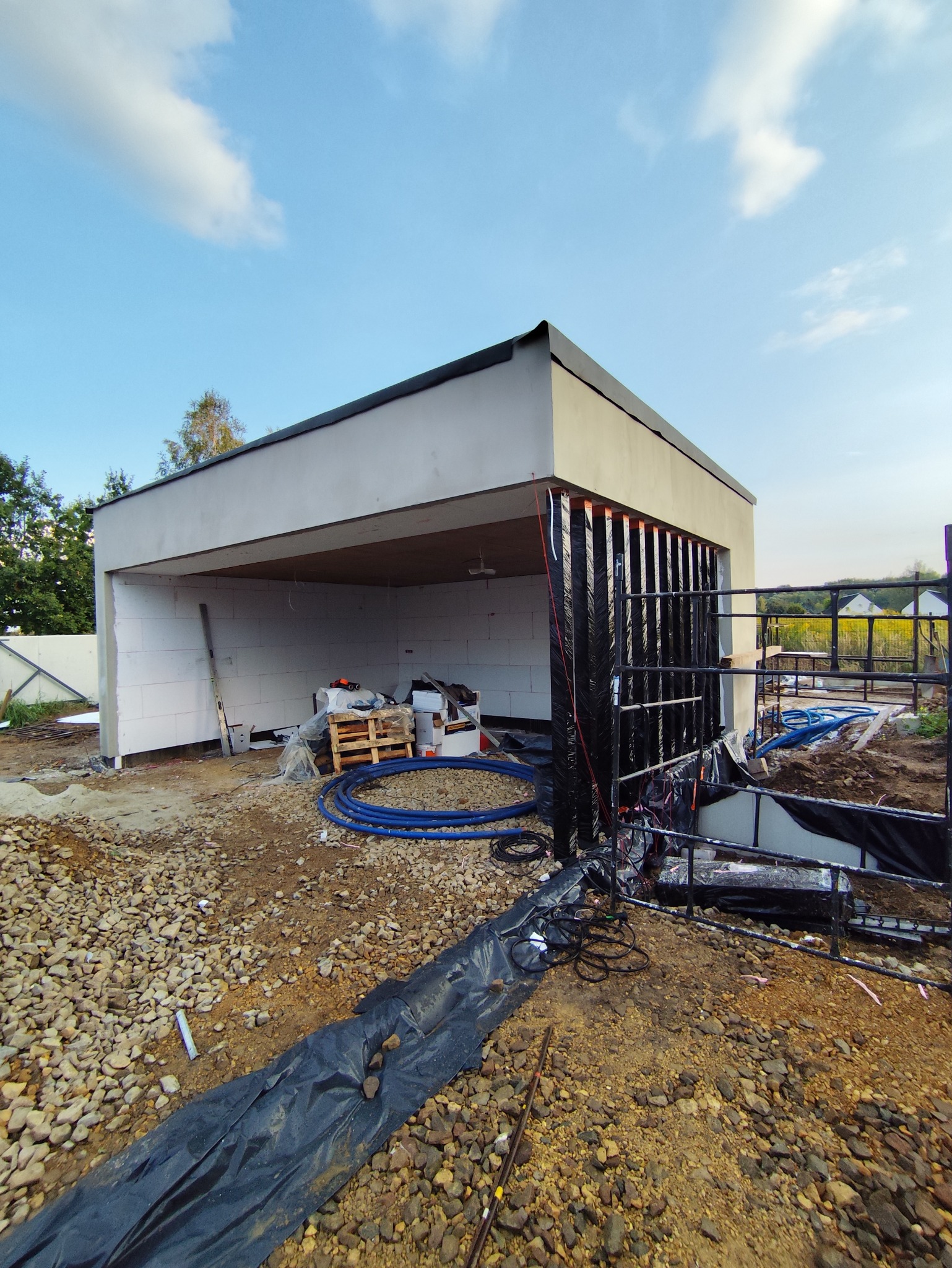 Nowoczesny garaż w trakcie budowy: ściany z betonu komórkowego, przygotowane zbrojenie. Widoczne materiały budowlane i narzędzia. Słoneczny dzień, błękitne niebo.