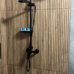 Usługi ogólnobudowlane Denis Janiak - Nowoczesna kabina prysznicowa z czarną armaturą na tle ściany z drewnianych paneli. Na półce kolorowa gąbka. Minimalistyczny design.
