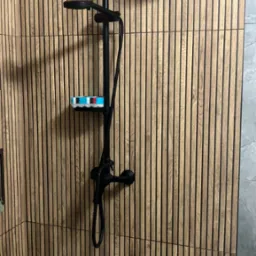 Nowoczesna kabina prysznicowa z czarną armaturą na tle ściany z drewnianych paneli. Na półce kolorowa gąbka. Minimalistyczny design.