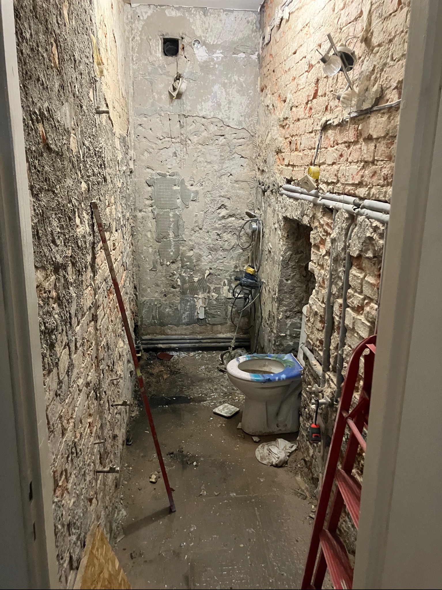 Wnętrze łazienki w trakcie demontażu: odsłonięte cegły, instalacje, usunięte płytki. Widoczny sedes, drabina i narzędzia. Przygotowanie do remontu z surowym wykończeniem.