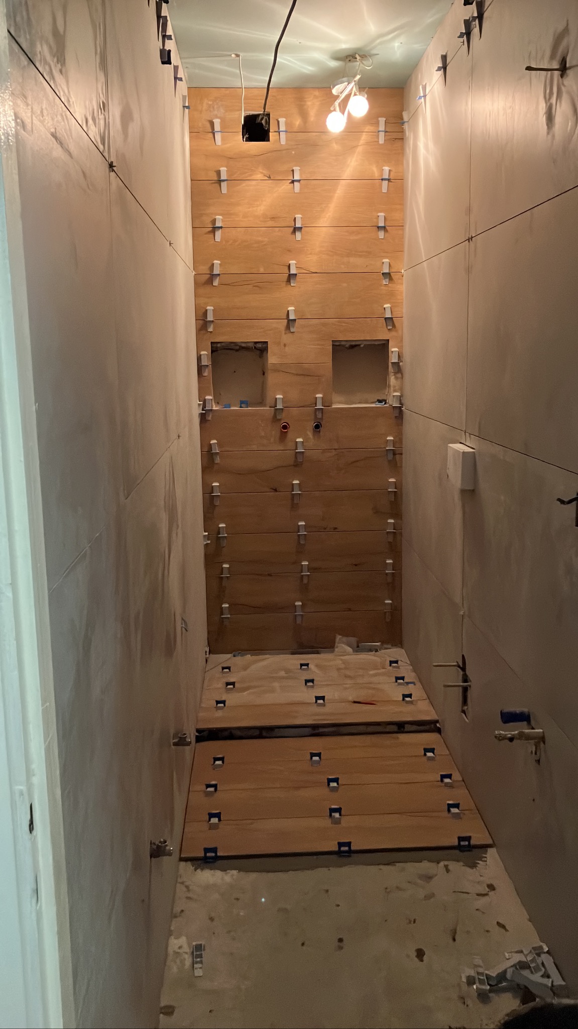Wąska łazienka w trakcie montażu drewnopodobnych płytek. Widoczne krzyżyki dystansowe, poziomice laserowe i wyprowadzone instalacje. Świecące żarówki na suficie.