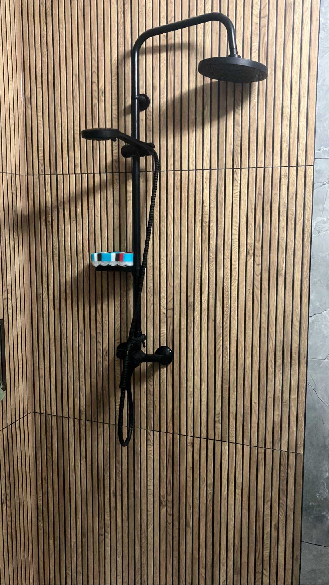 Nowoczesna kabina prysznicowa z czarną armaturą na tle ściany z drewnianych paneli. Na półce kolorowa gąbka. Minimalistyczny design.