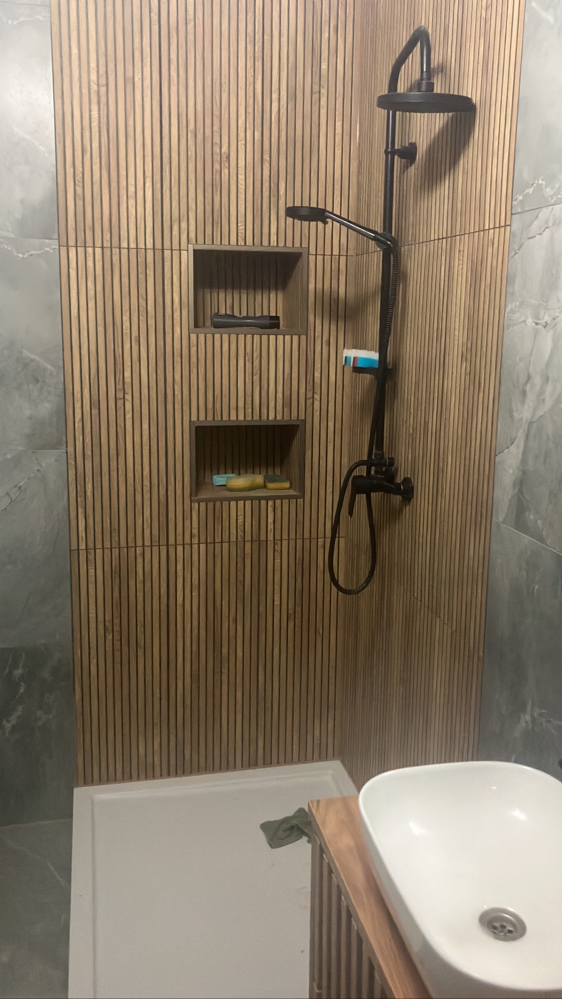 Nowoczesna łazienka z kabiną prysznicową wykończoną drewnopodobnymi panelami z wnękami na akcesoria, czarna armatura i umywalka nablatowa.
