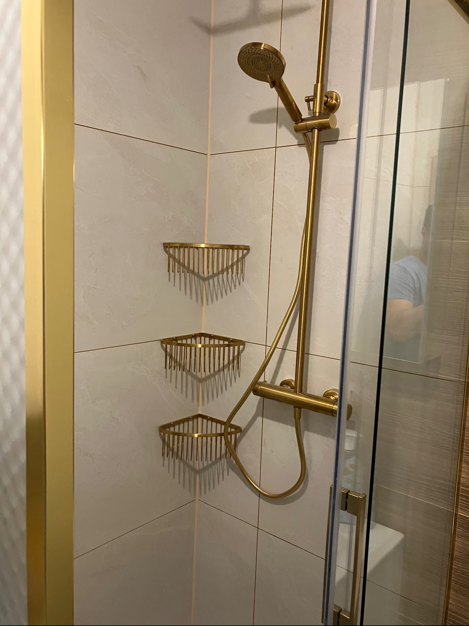 Wykończona kabina prysznicowa z złocistą armaturą: deszczownica, półki narożne. Białe płytki imitujące marmur. Szklane drzwi kabiny z złotymi detalami.