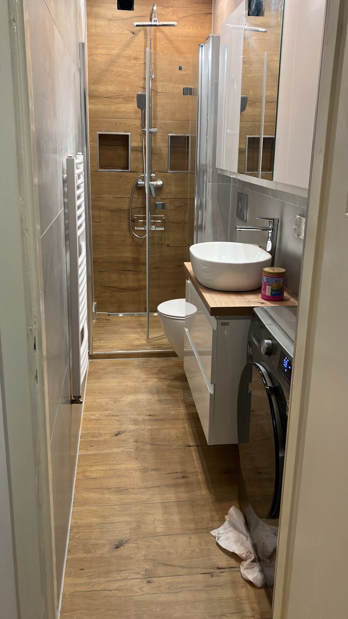 Nowoczesna łazienka z drewnopodobnymi płytkami, kabiną prysznicową typu walk-in, białą umywalką nablatową i pralką. Minimalistyczny design, ciepłe oświetlenie.