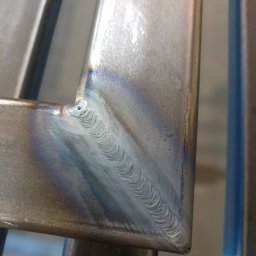 Nikstal - Zbliżenie na spawany narożnik metalowej konstrukcji. Widoczny precyzyjny spaw z charakterystycznym wzorem. Szlifowana powierzchnia metalu odbija światło.