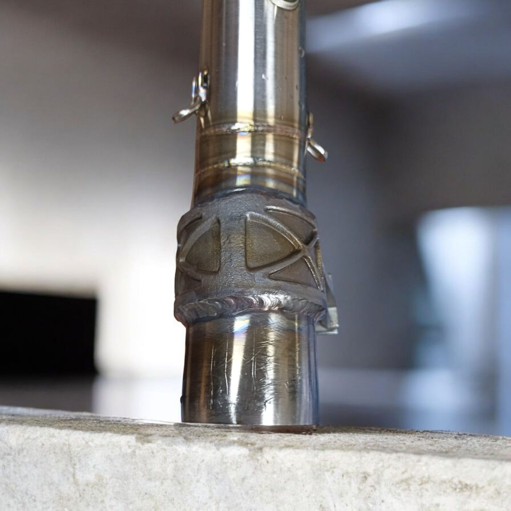 Szlifowany, spawany element metalowy z dekoracyjnym wzorem, widoczne ślady obróbki i spawania, osadzony na betonowej powierzchni, z bliska.