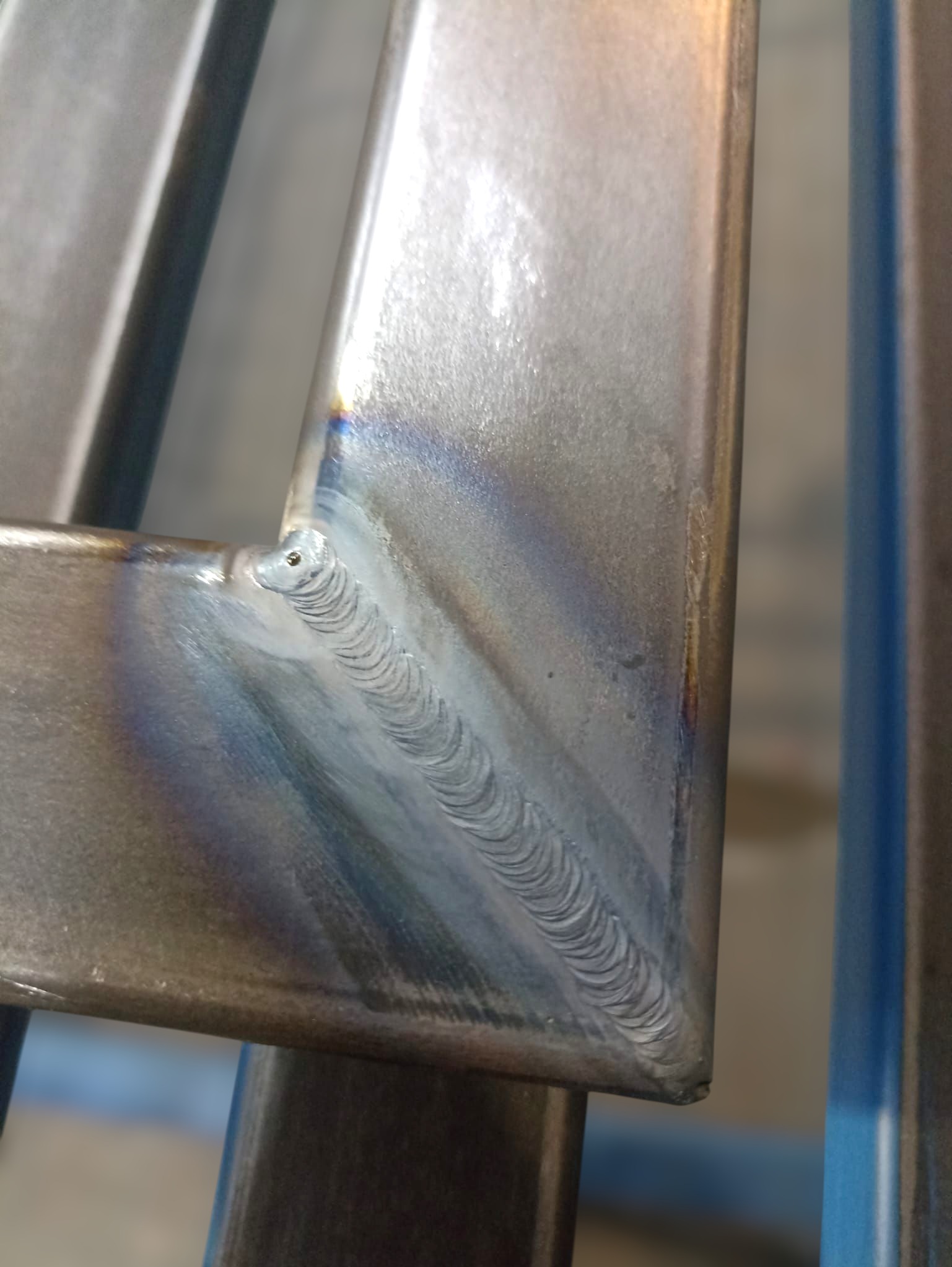 Zbliżenie na spawany narożnik metalowej konstrukcji. Widoczny precyzyjny spaw z charakterystycznym wzorem. Szlifowana powierzchnia metalu odbija światło.