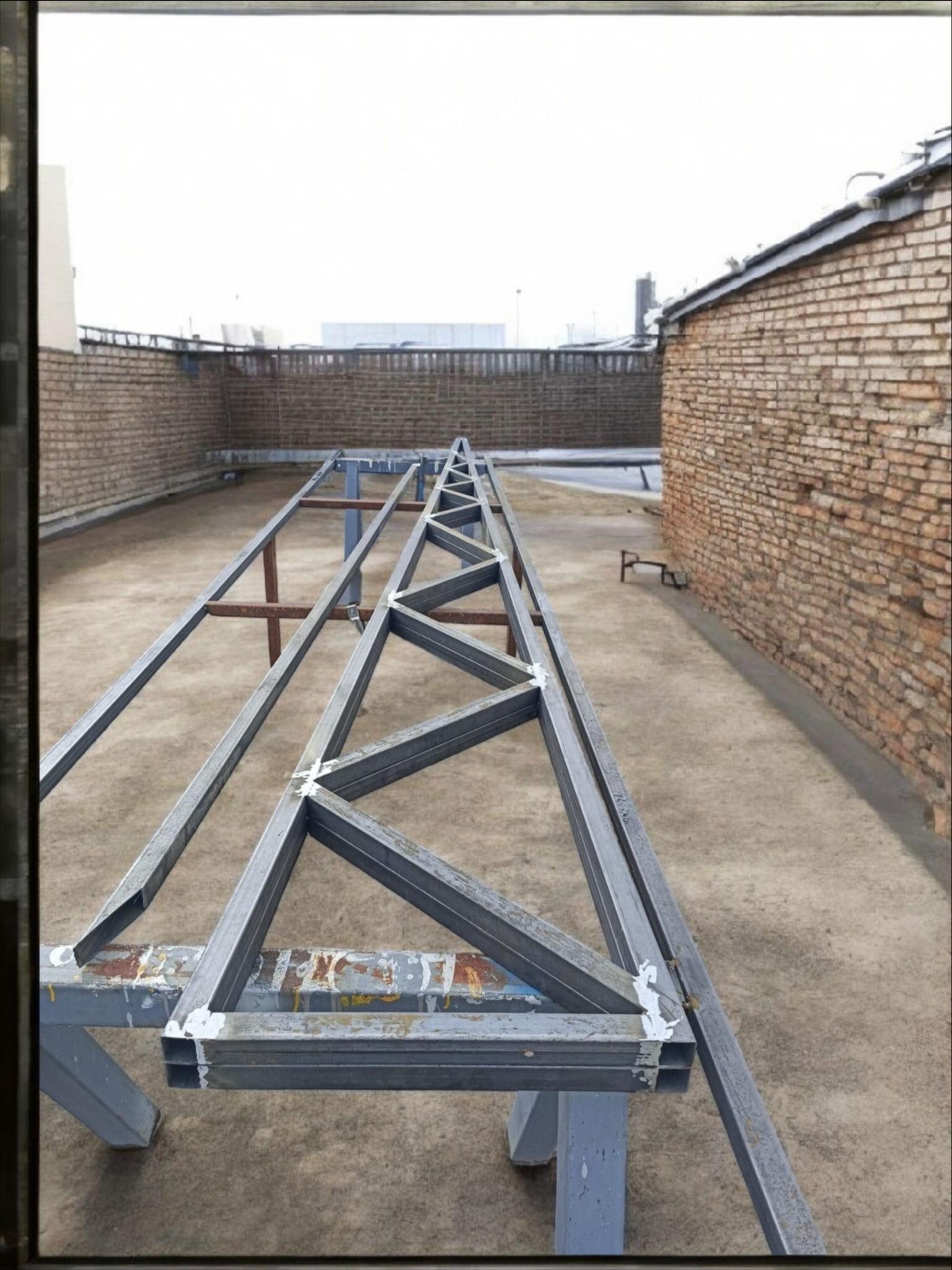 Metalowa konstrukcja spawana, prawdopodobnie fragment ogrodzenia lub balustrady, na tle ceglanego muru i szarej nawierzchni. Widoczne spawy i ślady po farbie.