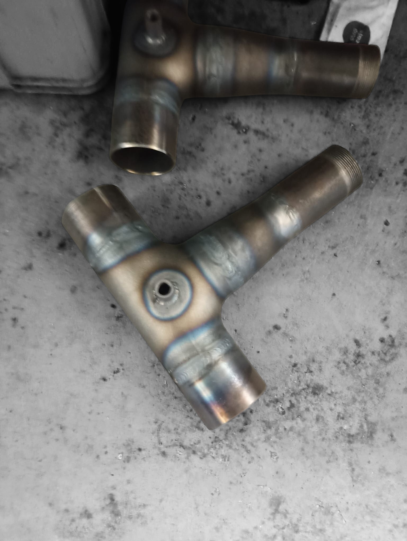 Dwa spawane, metalowe trójniki z widocznymi śladami spawania i otworami, leżące na szarej, betonowej powierzchni. Górny trójnik częściowo poza kadrem.