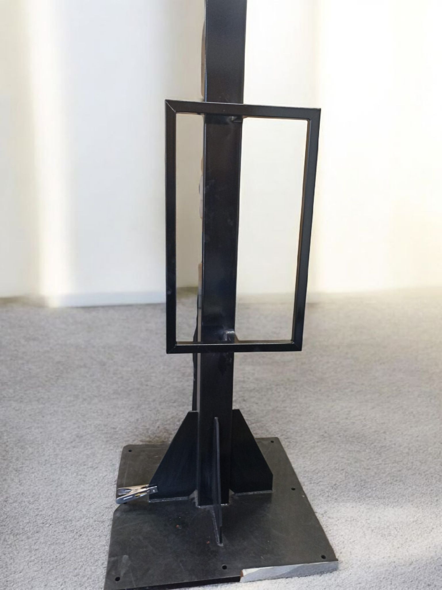 Czarna, metalowa konstrukcja z kwadratową ramą, stojąca na szarej wykładzinie, na tle białej ściany. Widoczne ślady spawania i mocowania.