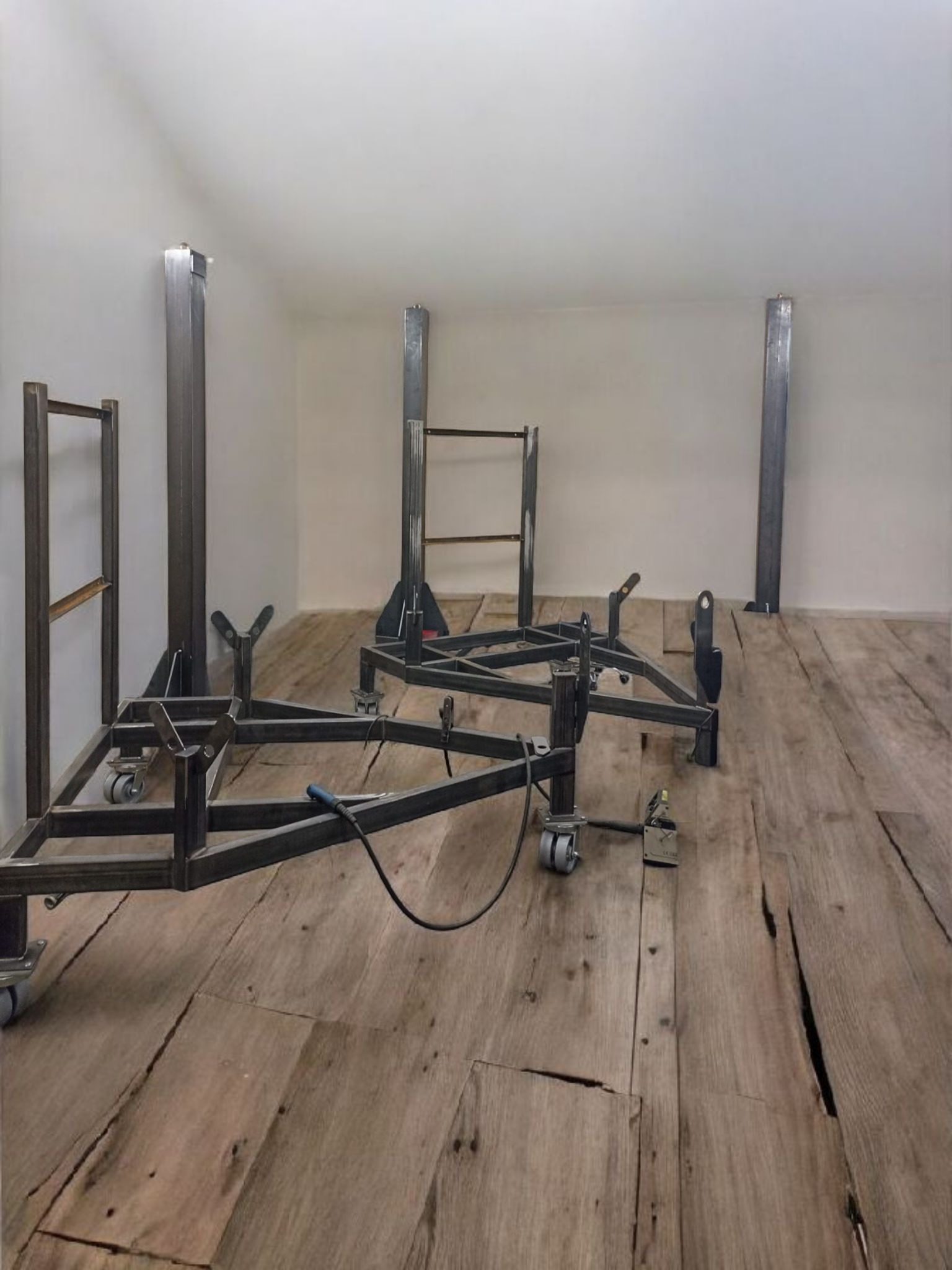 Wnętrze z surowymi, metalowymi konstrukcjami na kółkach i słupami, prawdopodobnie w trakcie montażu. Podłoga z desek o wyraźnej fakturze. Minimalistyczne, jasne ściany.