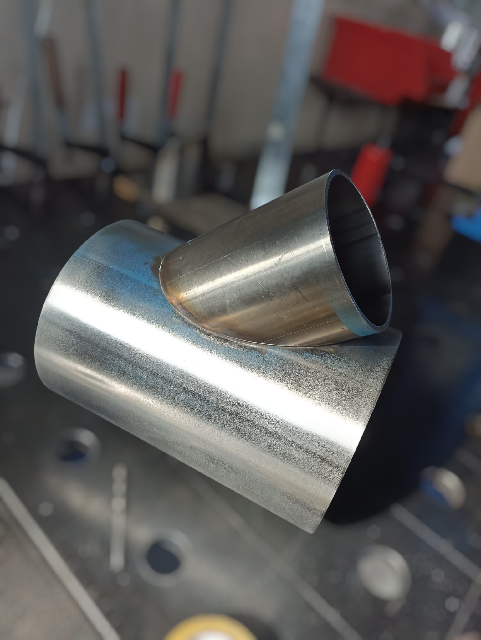 Precyzyjnie zespawane, nierdzewne rury pod kątem, widoczne ślady spawania. Metalowa powierzchnia z delikatnym połyskiem. Tło warsztatowe, narzędzia w tle.