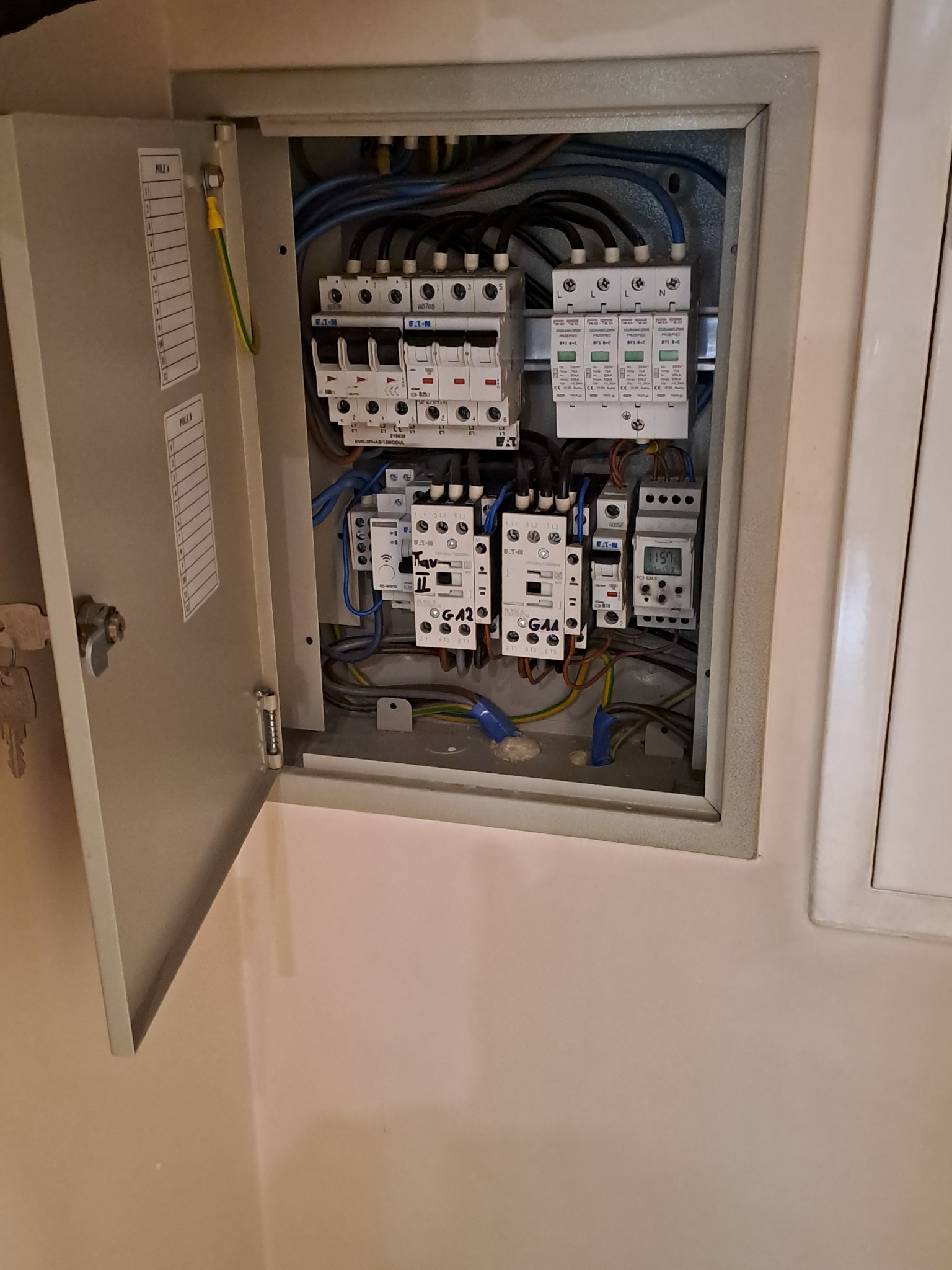 Otwarta skrzynka elektryczna z bezpiecznikami, przewodami i elementami sterującymi. Widoczne oznaczenia na urządzeniach i listwa z opisem obwodów na drzwiach.