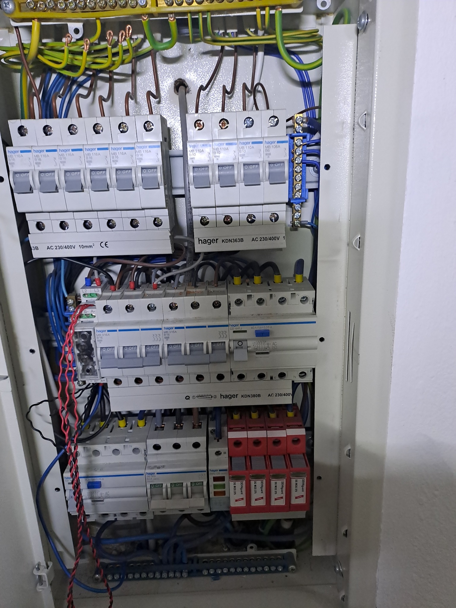 Otwarta skrzynka elektryczna z bezpiecznikami i przewodami. Widoczne elementy instalacji elektrycznej, w tym zabezpieczenia przeciwprzepięciowe i wyłączniki nadprądowe.