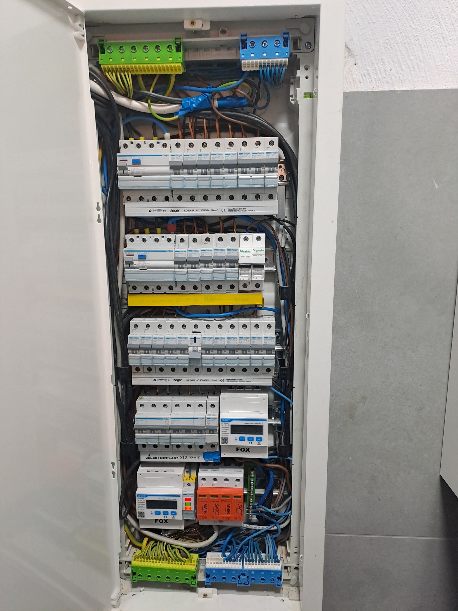 Otwarta skrzynka elektryczna z bezpiecznikami, przewodami i licznikami. Widoczne elementy marki Hager, Schneider i Elektro-Plast. Uporządkowany układ komponentów.