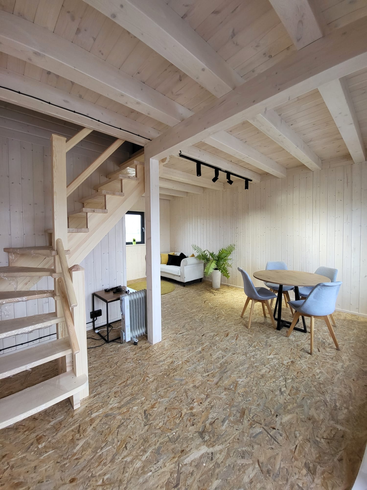 Jasne, drewniane wnętrze domu z surowymi schodami zabezpieczonymi folią, okrągłym stołem i sofą w tle. Minimalistyczny design z elementami naturalnego drewna.