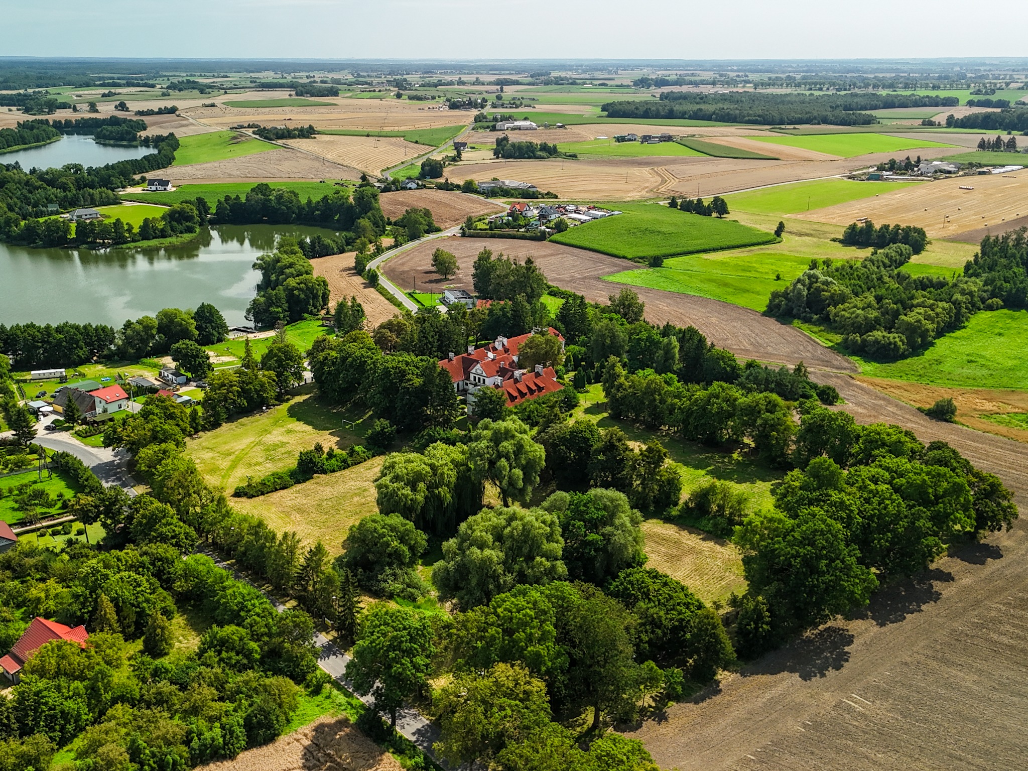 Rozległy krajobraz z widokiem z lotu ptaka na jezioro, pola uprawne i zabudowania, w tym dwór z czerwoną dachówką, otoczony zielenią drzew.