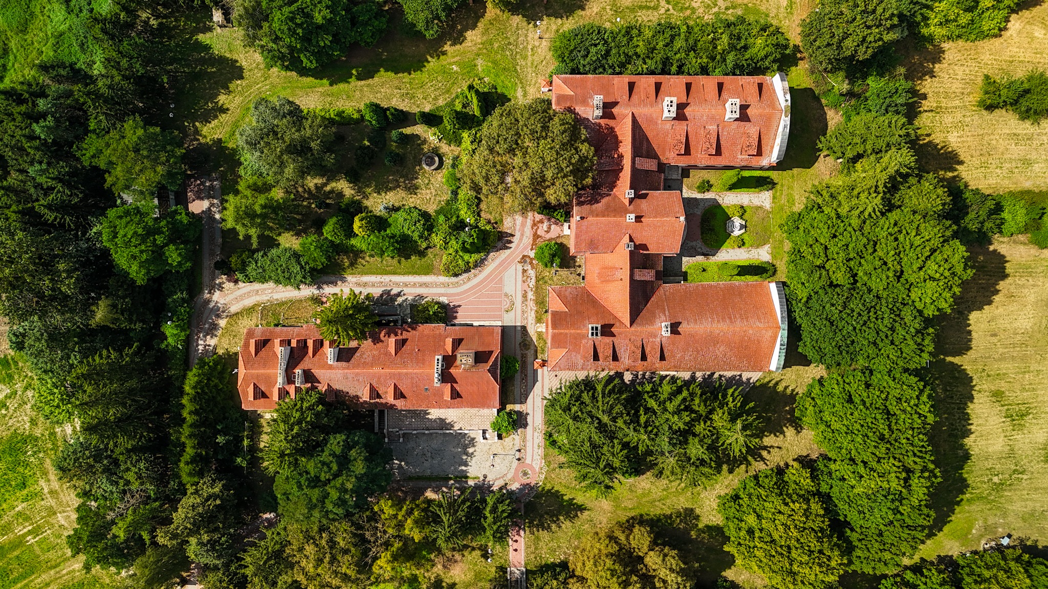 Widok z drona na rozległy kompleks budynków z czerwonymi dachami, otoczony zielenią drzew i trawników. Symetryczny układ budynków i ścieżek.