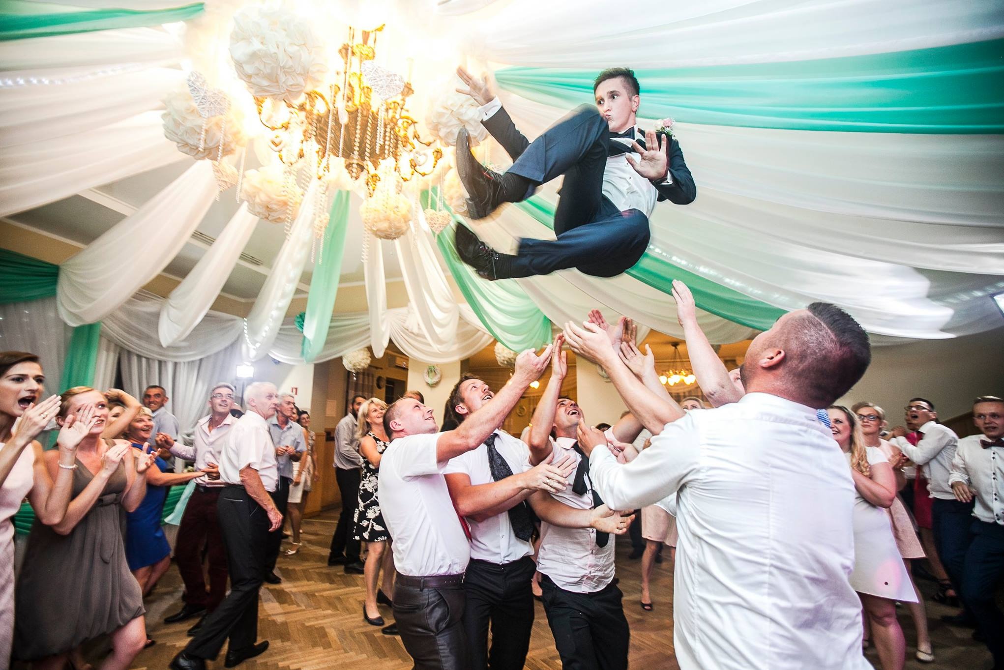 Uczestnicy wesela podrzucają pana młodego w garniturze. Sala udekorowana białymi i zielonymi tkaninami, w tle żyrandol. Radosna atmosfera.