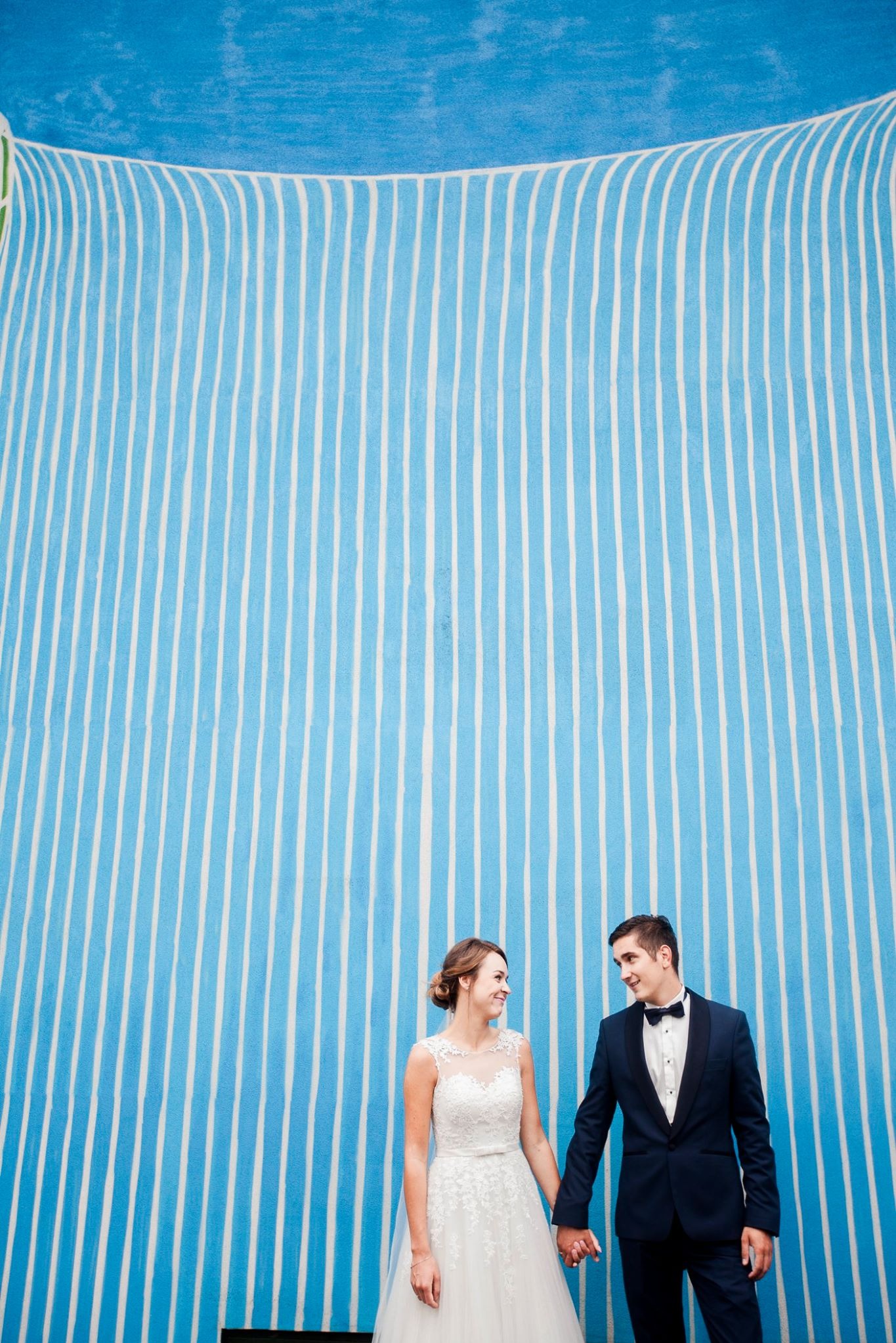 Para młoda trzymająca się za ręce na tle niebieskiej ściany w Wilamowicach. Panna młoda w białej sukni, pan młody w granatowym smokingu. Uśmiechnięci, patrzą na siebie.