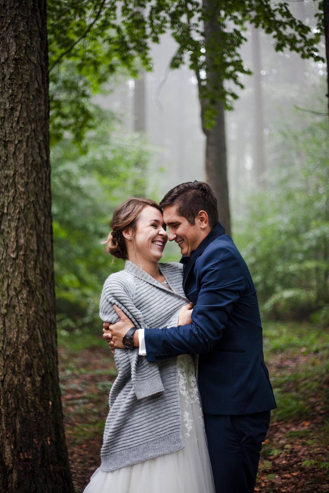Uśmiechnięta para w leśnej scenerii: Panna młoda w swetrze i sukni, pan młody w granatowym garniturze, blisko siebie, w tle drzewa i mgła. Romantyczna atmosfera.