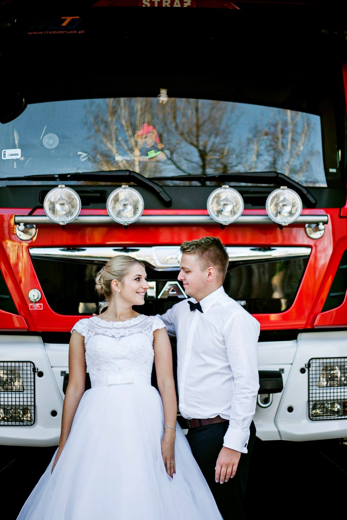 Uśmiechnięta para młoda, panna młoda w białej sukni i pan młody w koszuli z muchą, pozują na tle czerwono-białego wozu strażackiego z pluszową zabawką w kabinie.