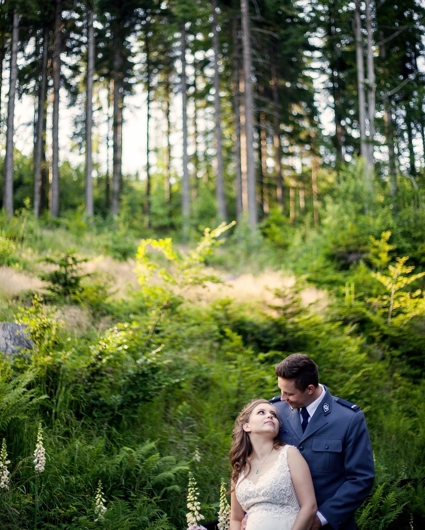Para w leśnej scenerii: kobieta w koronkowej sukni i mężczyzna w galowym mundurze, spoglądają na siebie z czułością, otoczeni zielenią drzew i paproci.
