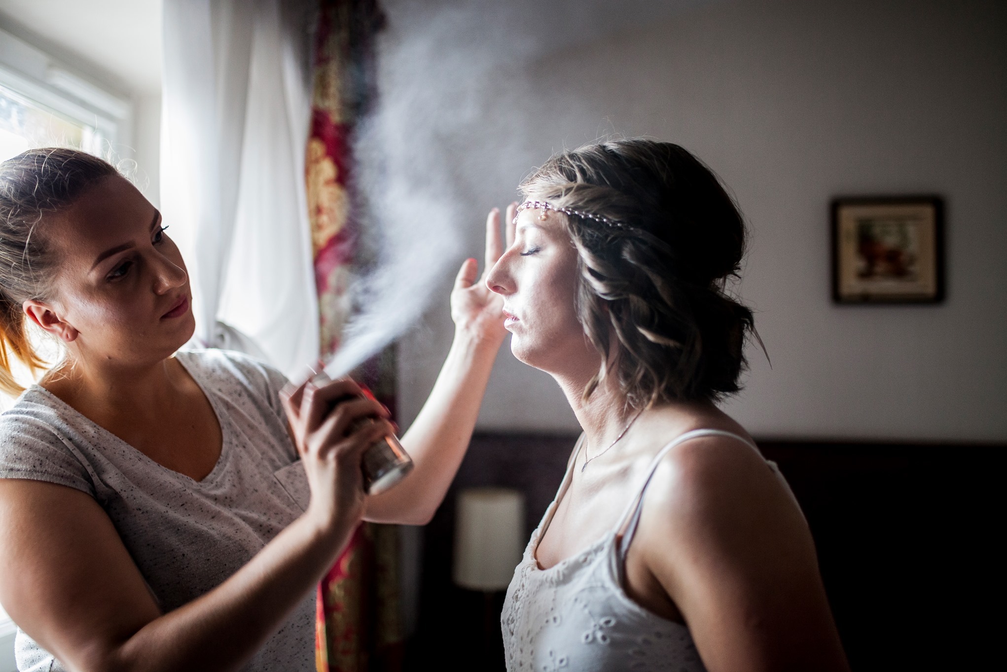 Makijażystka aplikuje utrwalacz na twarz panny młodej w białej sukience z ozdobą na głowie, przygotowując ją do sesji ślubnej w pokoju z oknem.