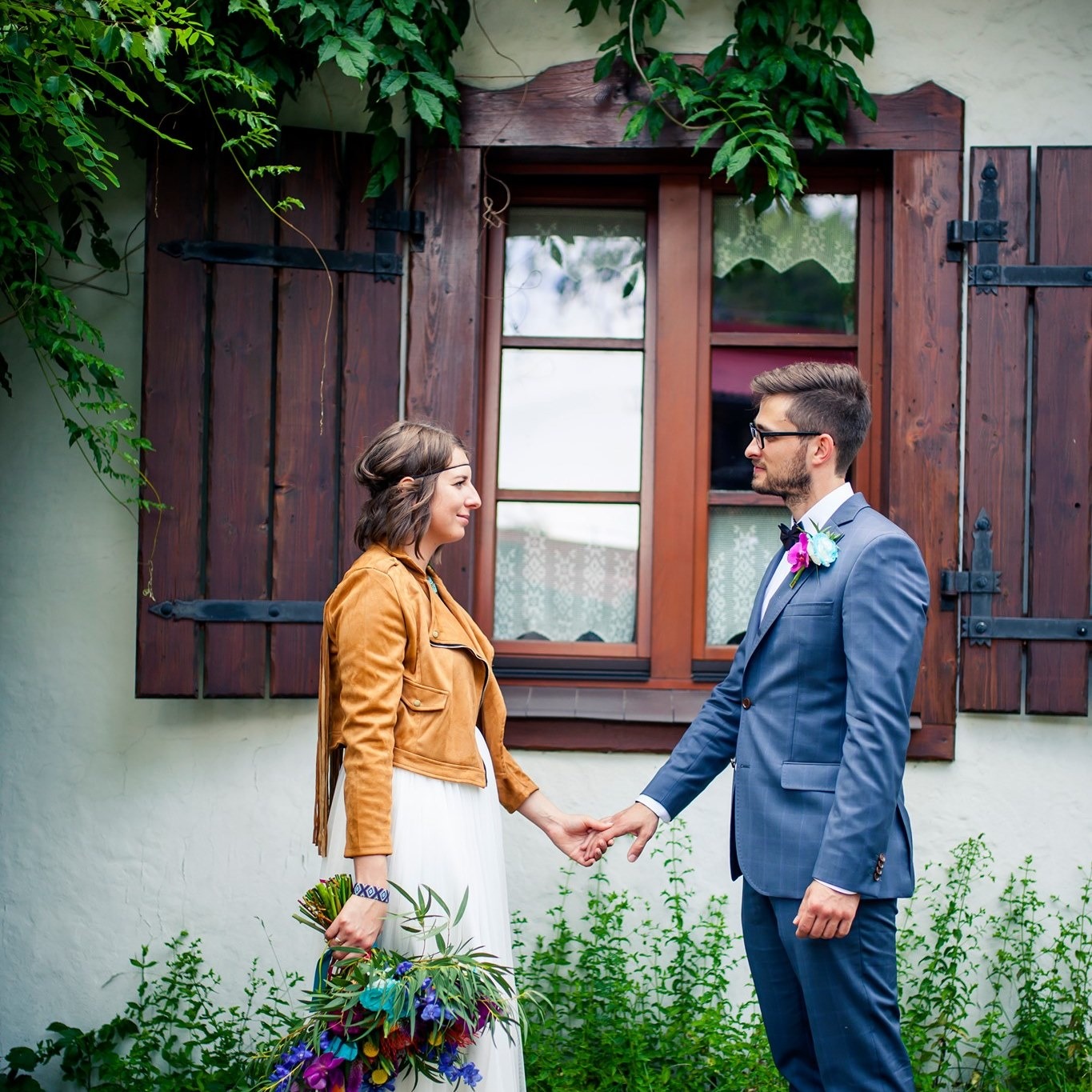 Para młoda trzymająca się za ręce na tle rustykalnego okna z okiennicami i zieleni. Bukiet kwiatów w ręku panny młodej, stylizacja boho.