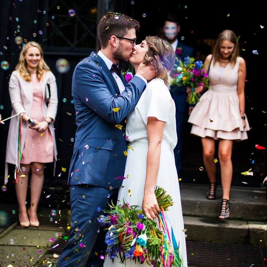 Młoda para w eleganckich strojach całuje się na tle kościoła, otoczona bańkami mydlanymi i konfetti, z gośćmi weselnymi w tle.