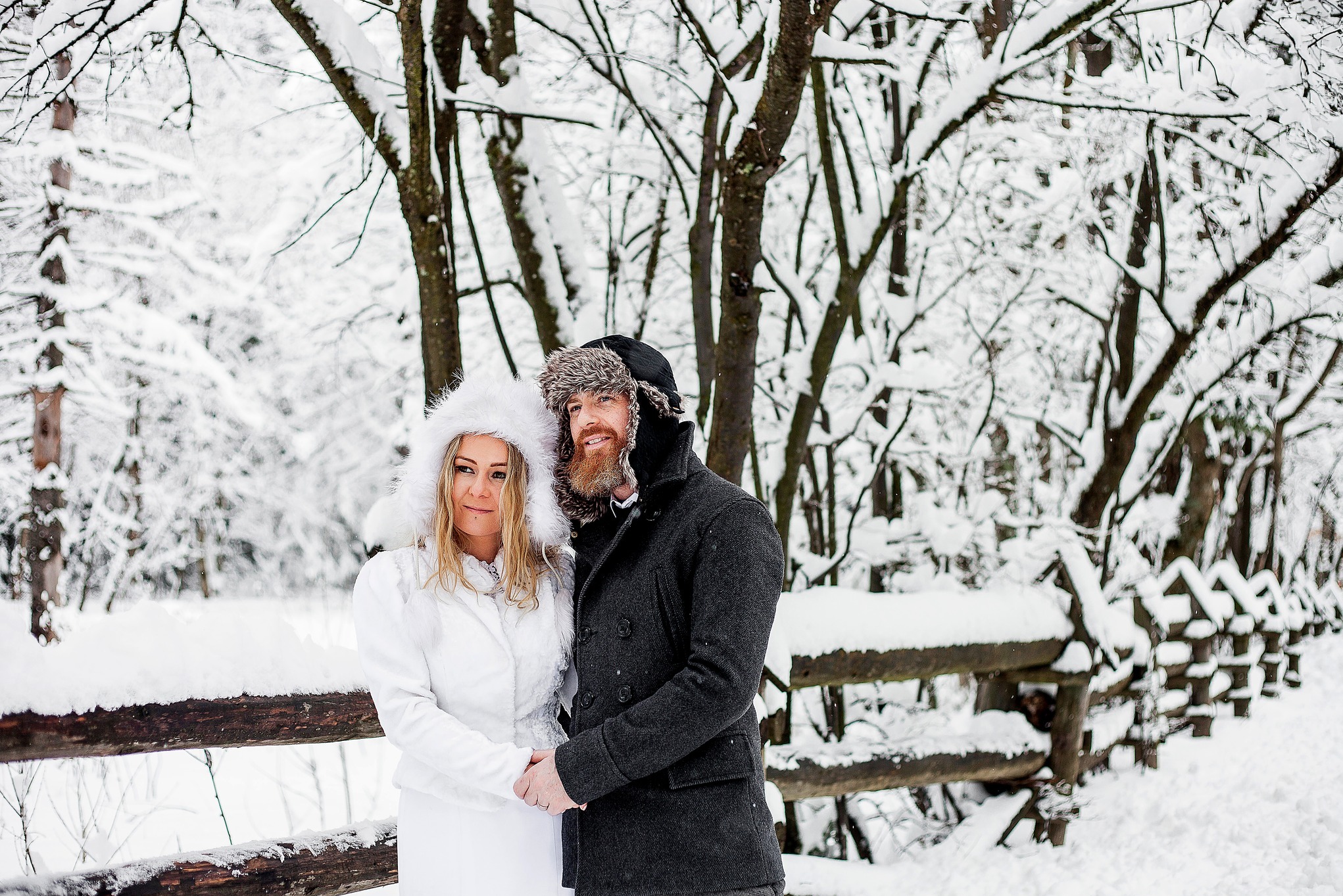 Para młoda trzymająca się za ręce na tle zimowego krajobrazu. Kobieta w białym futrzanym płaszczu i kapturze, mężczyzna w ciemnym płaszczu i futrzanej czapce. Śnieżny las i drewniane ogrodzenie.