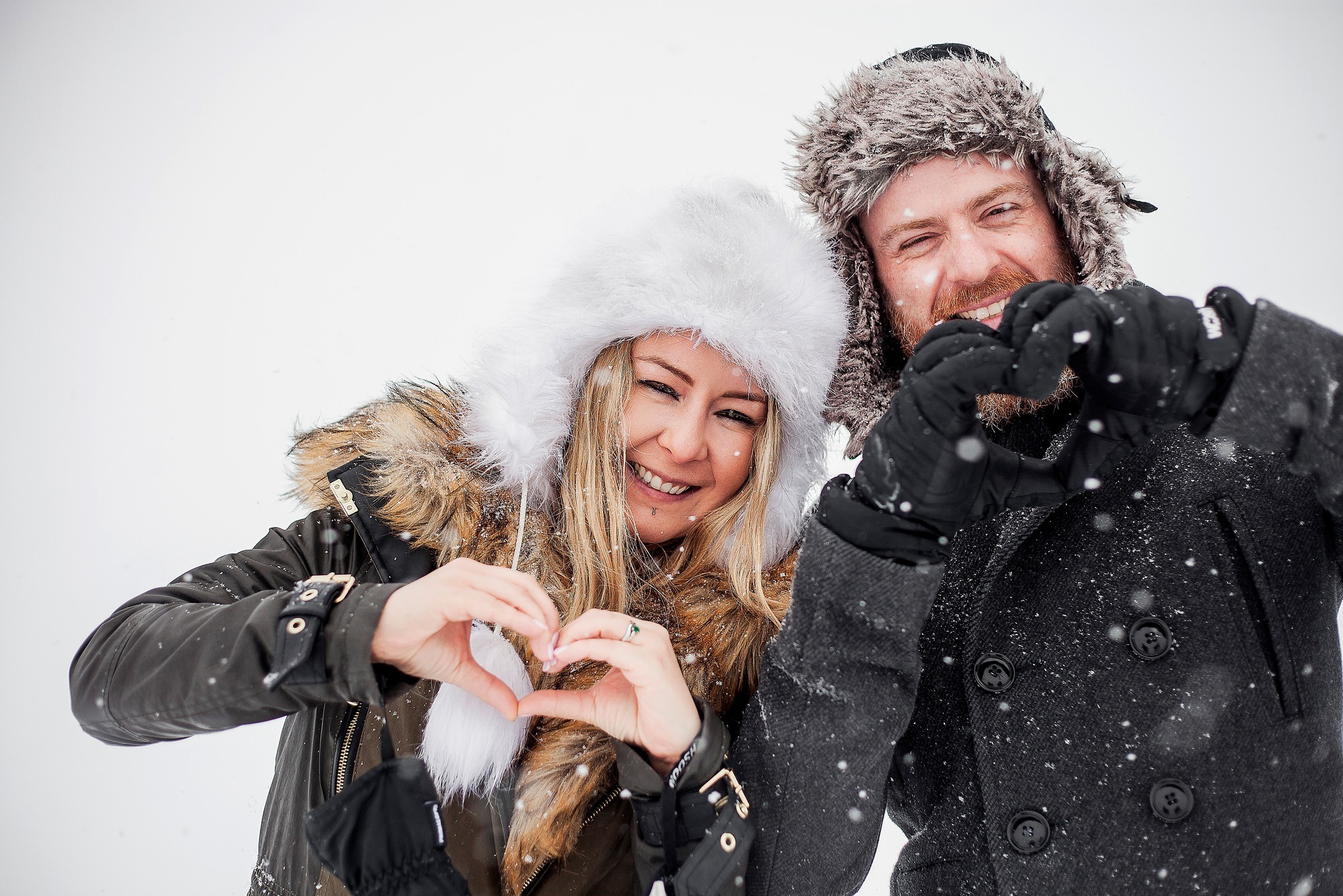 Uśmiechnięta para w zimowych ubraniach na tle śniegu, dłońmi formują serca. Kobieta w białej czapce z futerkiem, mężczyzna w uszatce. Padający śnieg.