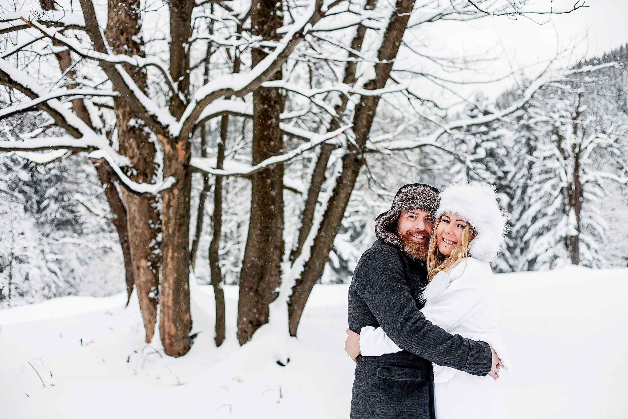 Uśmiechnięta para w zimowych strojach, obejmująca się na tle ośnieżonego lasu. Mężczyzna w futrzanej czapce, kobieta w białym futrzanym kapeluszu i płaszczu.