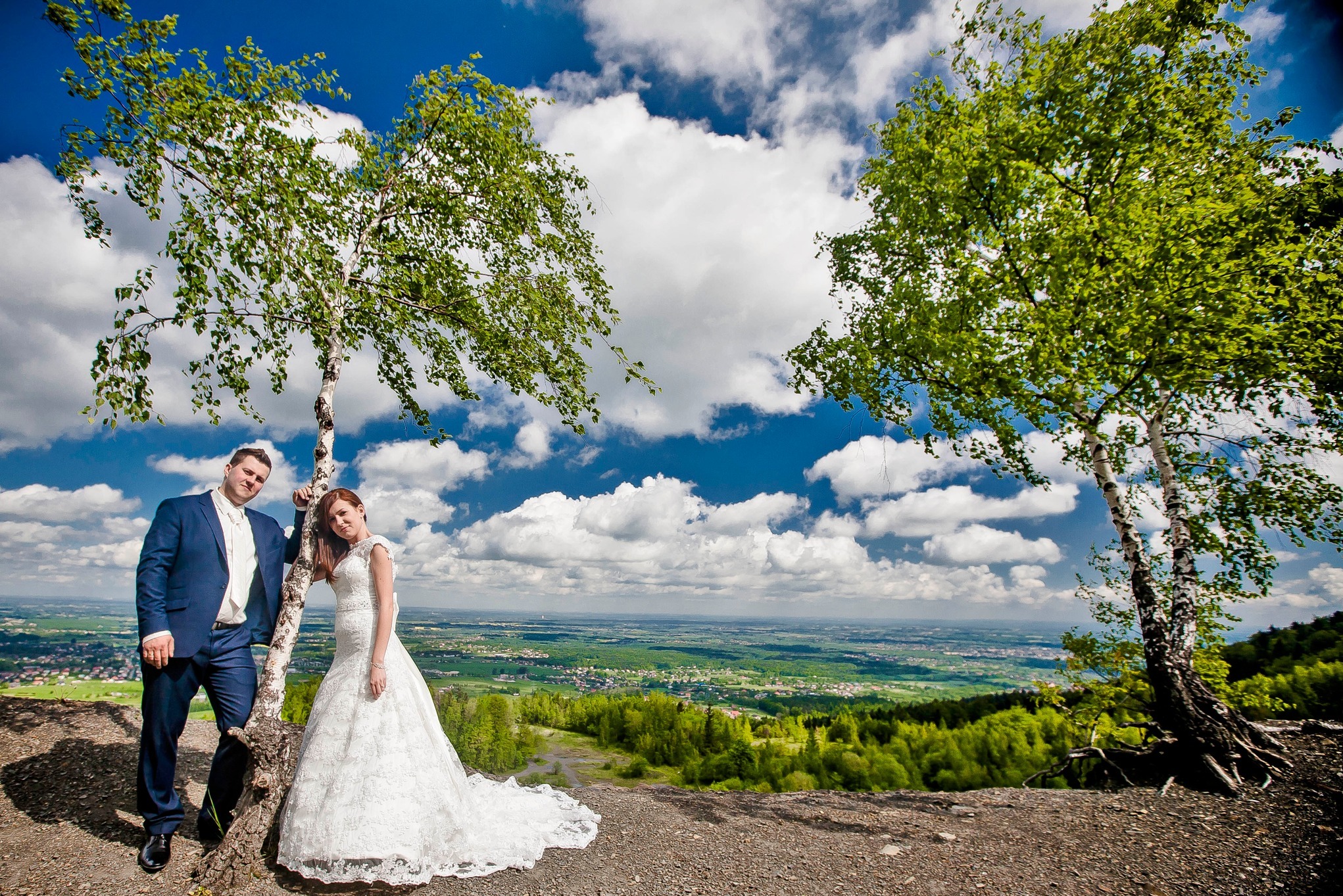 Para młoda pozuje oparta o brzozę na tle rozległego krajobrazu z błękitnym niebem i obłokami. Pan młody w granatowym garniturze, panna młoda w białej, koronkowej sukni.
