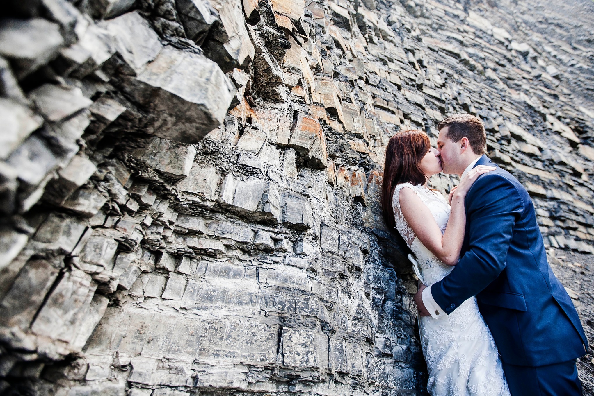 Pocałunek pary młodej na tle skalnej ściany. Ona w koronkowej sukni, on w granatowym garniturze. Nietypowa sceneria ślubna, surowy krajobraz.