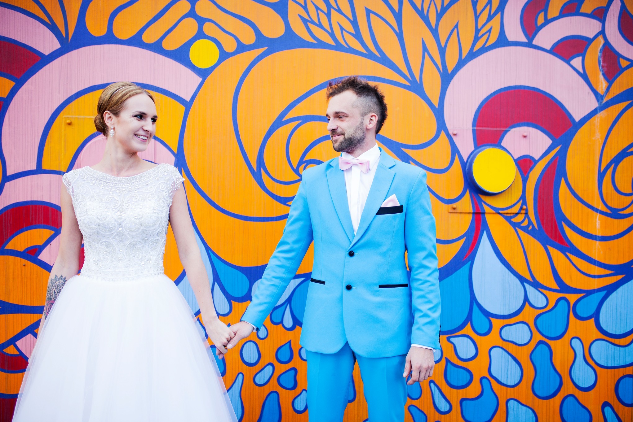 Para młoda trzymająca się za ręce na tle kolorowej grafiki. Panna młoda w białej sukni, pan młody w błękitnym garniturze z różową muchą. Uśmiechnięci, w plenerze.