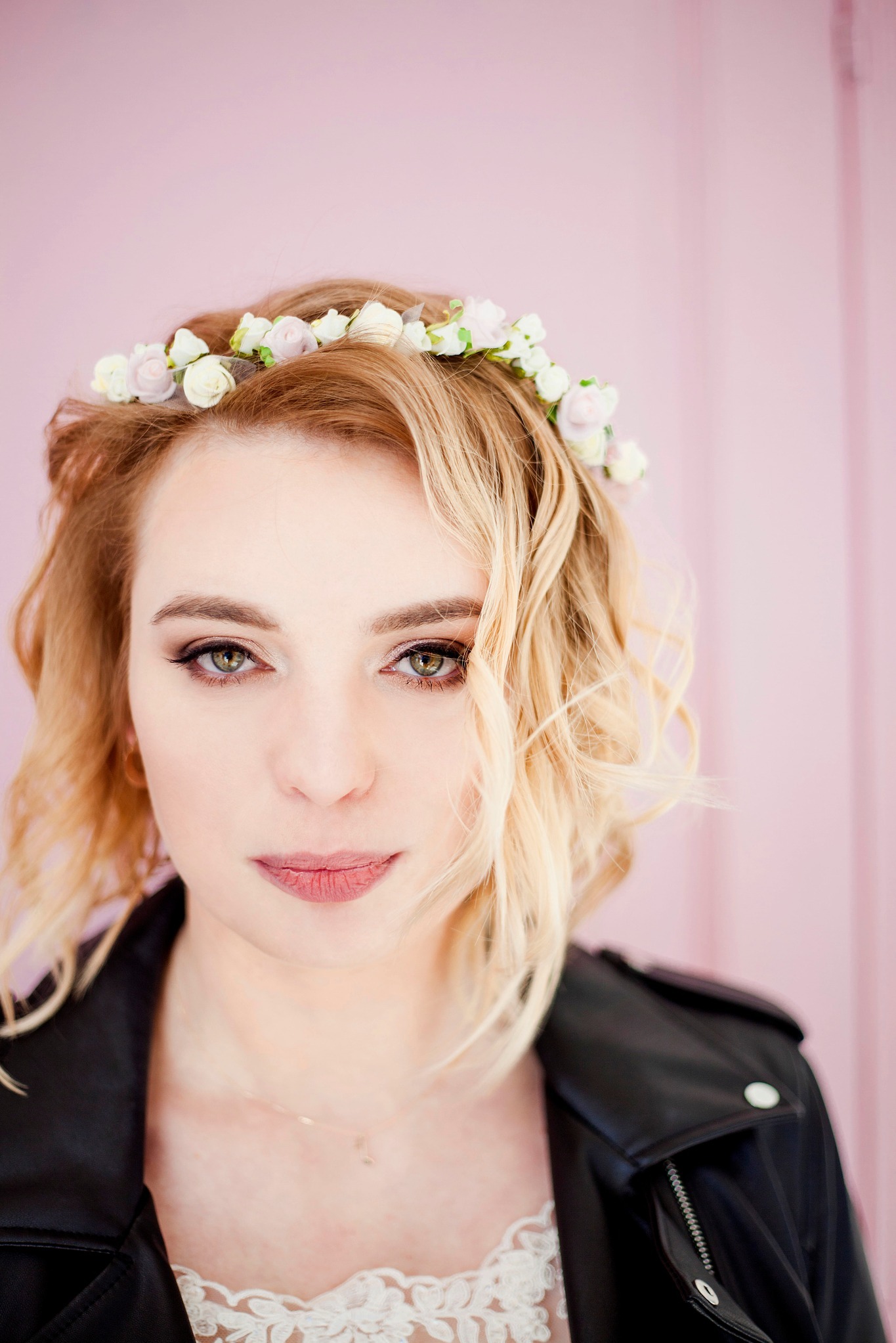 Portret kobiety z blond włosami, w wianku z białych kwiatów, ubrana w czarną skórzaną kurtkę na tle delikatnego, różowego tła.