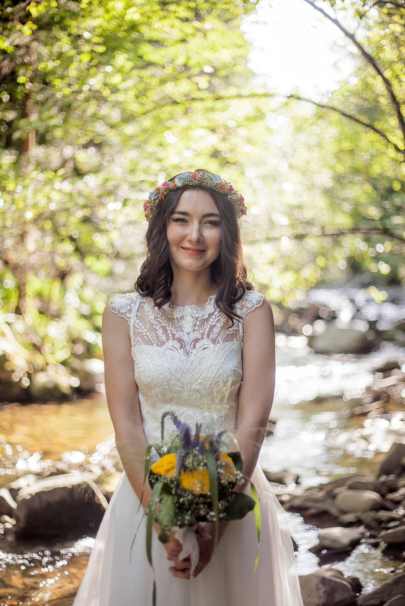 Uśmiechnięta panna młoda w koronie z kwiatów, w białej sukni z koronkową górą, trzyma bukiet na tle strumienia i zieleni. Romantyczna sceneria.